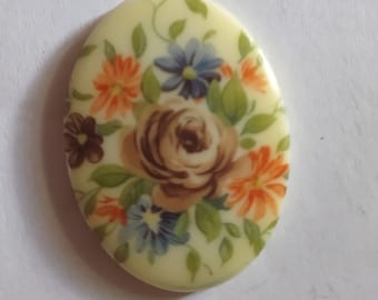May include: Oval-shaped, cream-colored ceramic pendant with a floral design. The design features a large brown rose with orange, blue, and green flowers surrounding it.