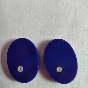 May include: A pair of oval-shaped earrings with a dark blue background and a small clear rhinestone in the center of each earring.