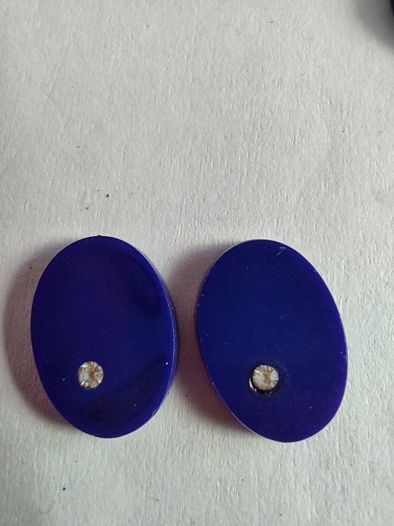 May include: A pair of oval-shaped earrings with a dark blue background and a small clear rhinestone in the center of each earring.