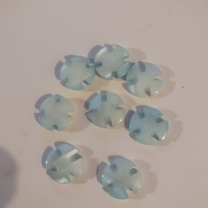 May include: A collection of eight light blue, fish-shaped beads. Each bead has a glossy finish and a cross-shaped design with rounded edges. The beads are scattered on a white surface, creating a delicate and aquatic aesthetic.