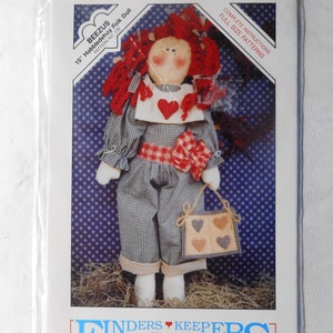 May include: A packaged craft pattern for a 15-inch Hobbledehoy Folk Doll by Beezus. The doll has red yarn hair, a heart-shaped detail, and a gingham outfit. The package includes full-size patterns and instructions. The Finders Keepers logo is at the bottom.
