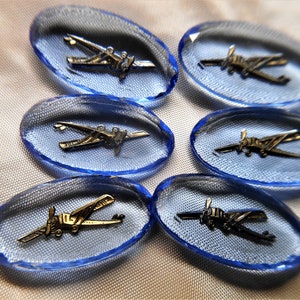 May include: Six blue glass oval-shaped buttons with a silver airplane design. The buttons are on a white fabric background.