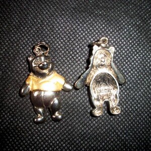 May include: Two Winnie the Pooh bear charms. One charm is silver with a gold-colored shirt. The other charm is silver.