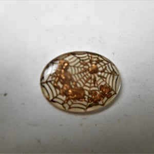 May include: A clear oval-shaped button with a gold spider web design and three spiders. The spiders are in the web and are facing different directions.