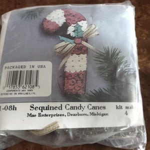 May include: A craft kit to make four sequined candy canes. The kit includes red, white, and green sequins, along with a white and gold ribbon. The packaging includes the text "Sequined Candy Canes" and "Mac Enterprises, Dearborn, Michigan".