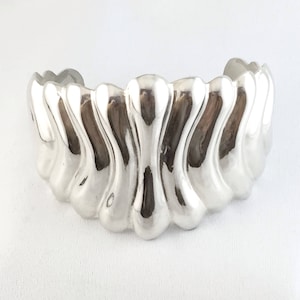 May include: A silver bracelet with a wavy, organic design. The bracelet is made of smooth, polished metal and has a rounded shape.
