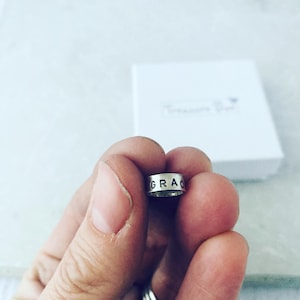May include: A small, cylindrical silver-toned metal bead featuring the word GRACE stamped in black capital letters along its outer surface. The bead has a smooth, polished finish and a hollow centre, designed for use as a jewellery component.