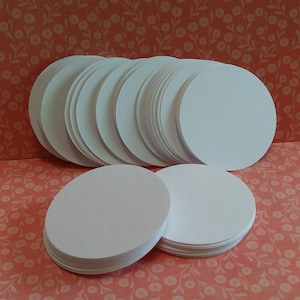 May include: A stack of white, circular paper cutouts. The cutouts are arranged in multiple stacks, with a few stacks in the foreground and a larger stack in the background. The paper appears to be smooth and uniform in color.