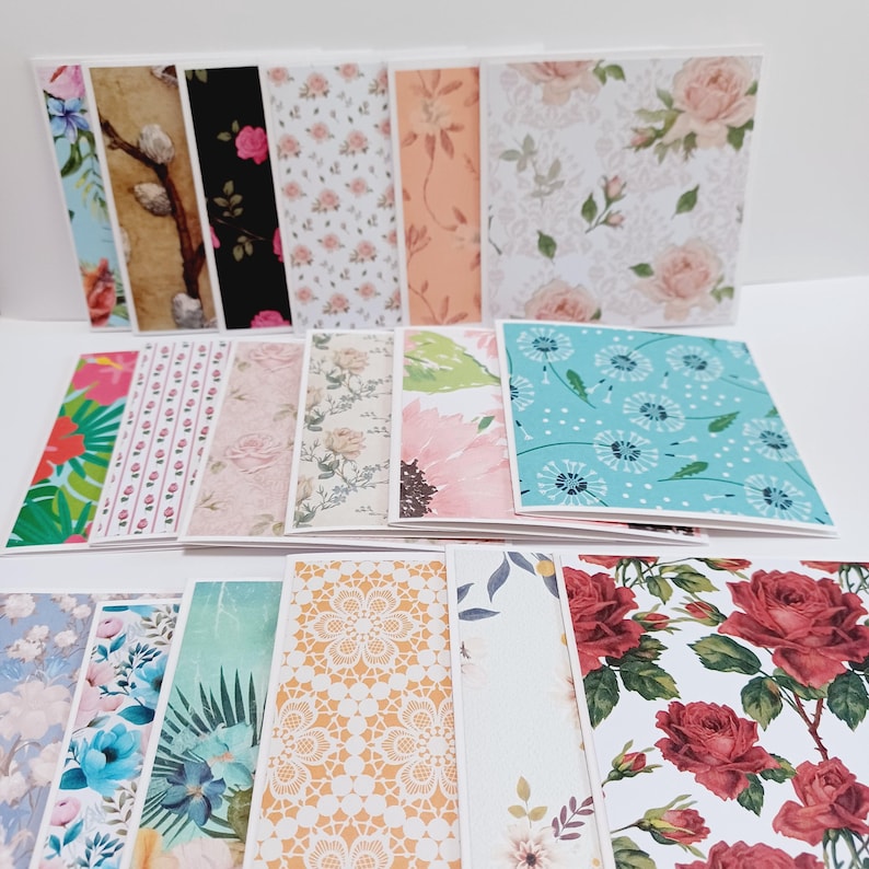 May include: A collection of folded greeting cards featuring a variety of printed patterns including floral designs with red roses, pink blossoms, and blue dandelions, as well as geometric lace motifs, tropical leaves, and botanical illustrations.