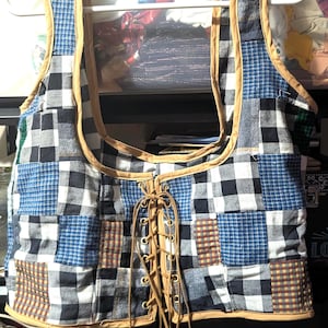 May include: A patchwork vest with a square neckline and a lace-up front. The vest features a variety of fabric patterns, including black and white checks, blue squares, and brown plaid. The edges are trimmed with a tan-colored material.
