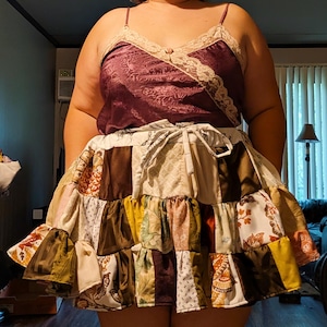 Op de afbeelding: Een donkerpaarse camisole met een V-hals, beige kanten rand en een klein roosje, gecombineerd met een veelkleurige patchworkrok bestaande uit diverse bruine, crèmekleurige en mosterdgele stoffen vierkanten met bloemen- en geometrische patronen.