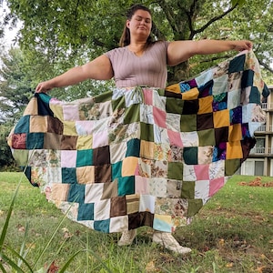 Könnte beinhalten: Ein bodenlanger Patchwork-Rock aus verschiedenen quadratischen Stoffstücken in Grün-, Braun-, Rosa-, Gelb- und Weißtönen, wobei einige Quadrate florale Muster aufweisen. Der Rock hat einen Kordelzug in der Taille und eine weite, ausgestellte Form.