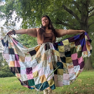 Könnte beinhalten: Ein bodenlanger Patchwork-Rock mit einer Vielzahl quadratischer Stoffflicken in Braun-, Rosa-, Gelb-, Grün- und Violetttönen, einschließlich floraler und einfarbiger Muster. Der Rock hat einen Kordelzug in der Taille und eine weite, ausgestellte Form.