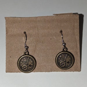 May include: A pair of gold-toned earrings with a clock face design. The clock face has Roman numerals and a gear design.