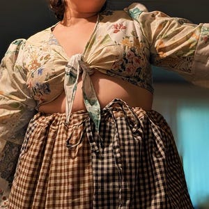 May include: A two-piece outfit featuring a floral print tie-front top and a checkered skirt. The top has long sleeves and a V-neckline. The skirt is a mix of brown and black checkered patterns, creating a unique visual contrast.