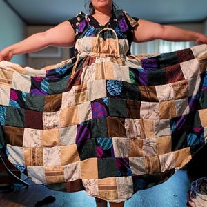 May include: A floor-length dress featuring a patchwork design composed of various square fabric panels in shades of tan, dark green, black, and purple, with some squares displaying floral patterns and others showing a blue and green plaid print.