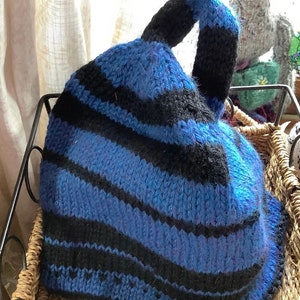 May include: A blue and black striped knitted hat with a loop handle. The hat is sitting in a wicker basket.