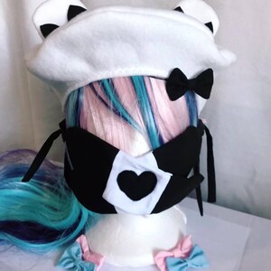 May include: White beret with black ears and a black bow. A black face mask with a white heart and black trim. Two pink and blue bows.