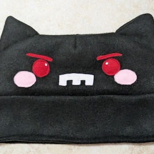 Puede incluir: Gorro negro con orejas de gato y una cara de dibujos animados. La cara presenta ojos rojos, mejillas rosas y una boca blanca. El sombrero está hecho de un material suave.