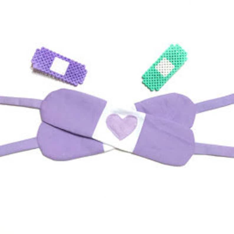 May include: Purple fabric eye mask with a white heart-shaped patch and two purple fabric straps. Two small purple and green beaded bandages are also shown.