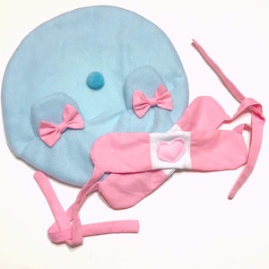 May include: A light blue plush hat with pink bows and a white heart on a pink headband. The hat has a small blue pom-pom on top.