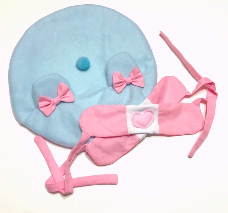 May include: A light blue plush hat with pink bows and a white heart on a pink headband. The hat has a small blue pom-pom on top.