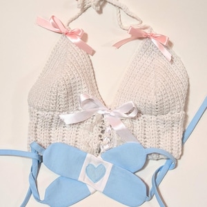 May include: A white crocheted halter top with pink ribbon ties and a white satin ribbon tie at the center. A blue fabric band with a white heart is included.