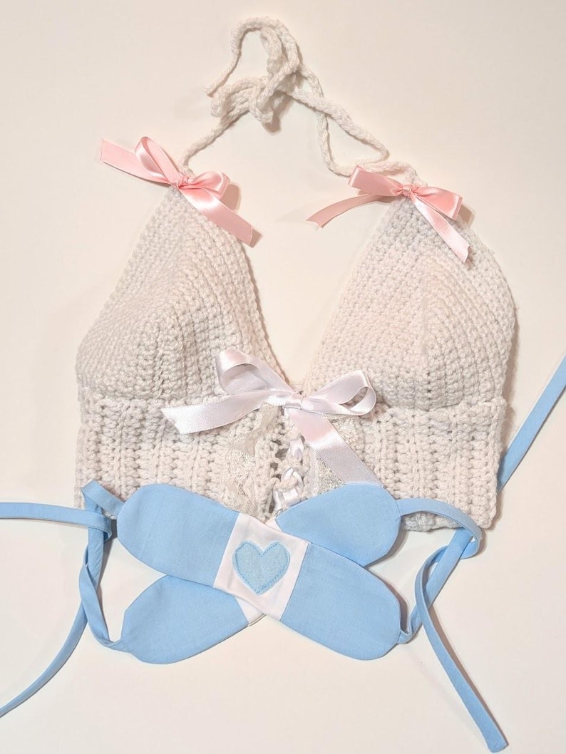 May include: A white crocheted halter top with pink ribbon ties and a white satin ribbon tie at the center. A blue fabric band with a white heart is included.