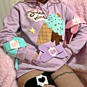May include: A lavender hoodie with a chocolate mint ice cream cone graphic and a star. The hoodie has a hood with a drawstring and long sleeves. The model is wearing the hoodie with a black fishnet top and a black heart-shaped patch on their thigh.