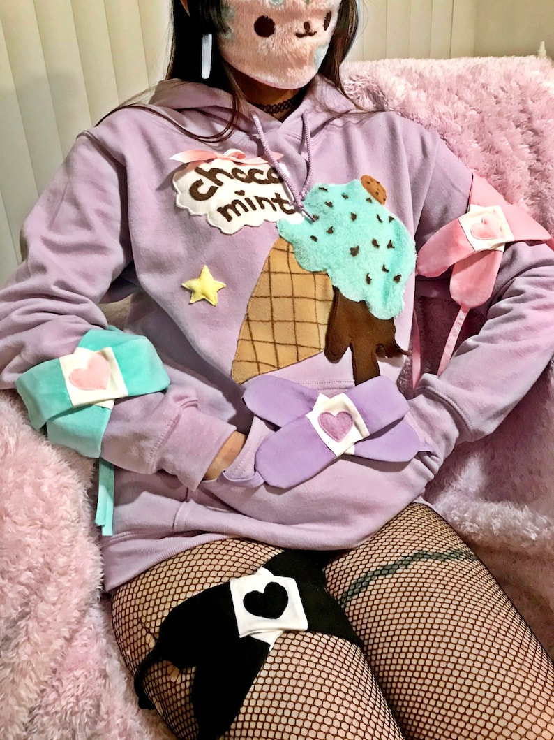 May include: A lavender hoodie with a chocolate mint ice cream cone graphic and a star. The hoodie has a hood with a drawstring and long sleeves. The model is wearing the hoodie with a black fishnet top and a black heart-shaped patch on their thigh.