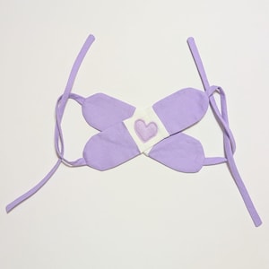 May include: A lavender fabric headband with a white band and a purple heart in the center. The headband has two long ties that can be tied at the back of the head.