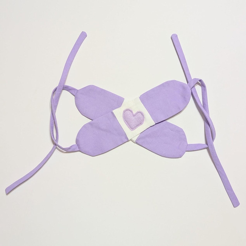 May include: A lavender fabric headband with a white band and a purple heart in the center. The headband has two long ties that can be tied at the back of the head.