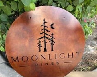 May include: A round, copper-colored metal sign with the words "MOONLIGHT PINES" cut out. The sign features a silhouette of pine trees and a crescent moon. Two small holes are at the top. The sign is displayed outdoors, with green foliage in the background.