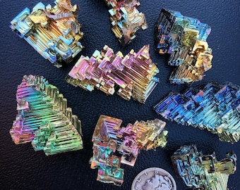 May include: A collection of iridescent bismuth crystals in various shapes and sizes. The crystals display a spectrum of colors, including blues, greens, yellows, and purples, with a metallic sheen. A silver coin is included for size reference.