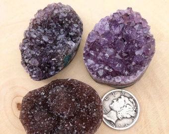 May include: Three amethyst crystal clusters, each with a different shape and size. The crystals are a deep purple color and have a natural, rough texture. A silver coin is included for scale.