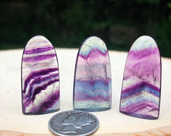 May include: Three polished fluorite stones with a rounded top and a flat base, displaying bands of purple, white, and blue hues. A silver coin is placed near the stones. The stones are displayed on a wooden surface.