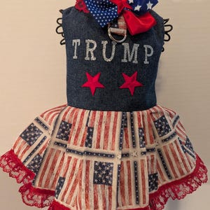 May include: A blue denim dog dress with a red, white, and blue bow, two red stars, and the word "TRUMP" in silver glitter. The skirt is made of a red, white, and blue American flag print fabric with a red lace trim.