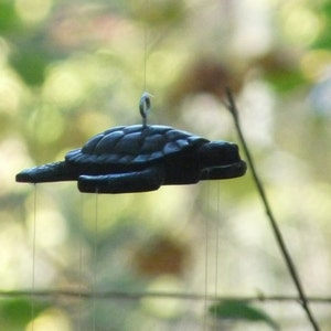 Puede incluir: Un carillón de viento de metal negro en forma de tortuga que cuelga de una cuerda. La tortuga está suspendida en el aire con cuerdas que cuelgan de su cuerpo.