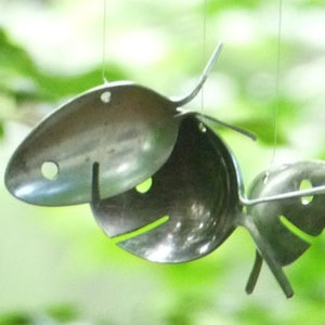 May include: Three silver spoons are suspended from the ceiling and are shaped like fish. The spoons are hanging against a green and white blurred background.