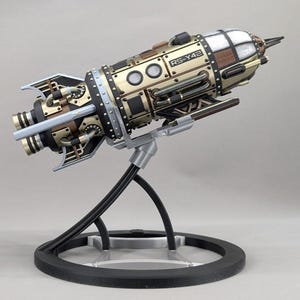 Puede incluir: Un modelo detallado de cohete espacial con acabado metálico dorado y negro, remaches visibles, tuberías y una ventana de cabina. El lateral del casco lleva el texto R5-Y42. El cohete está montado sobre un soporte circular negro y plateado.