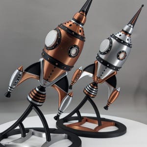以下が含まれることがあります： メタリックカッパーとメタリックシルバーの2つのロケットフィギュア。それぞれ黒いアクセント、尖ったノーズコーン、円形の窓、3つのフィンが特徴です。各フィンの側面にはRS-X94という文字が記されています。各ロケットは、サポート構造を備えた黒い円形のベースの上に立っています。