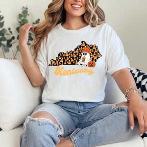 Puede incluir: Camiseta blanca con un gráfico de un fantasma sosteniendo una calabaza y maíz dulce en forma del estado de Kentucky. El texto "Kentucky" está escrito en una fuente retro debajo del gráfico.