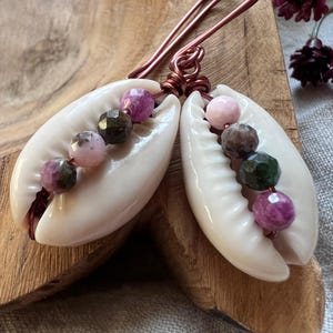 May include: A pair of handcrafted earrings featuring white cowrie shells and colorful gemstone beads. The earrings have copper wire accents and are displayed on a wooden surface.