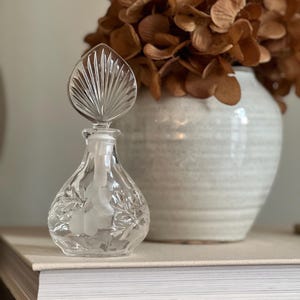 May include: A clear glass perfume bottle with a leaf-shaped stopper. The bottle has a pear-shaped body with etched floral designs. A white vase with brown dried flowers is in the background.