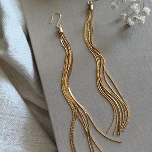May include: A pair of long gold-toned dangle earrings featuring multiple delicate, thin, flat-link chains that hang from a simple hook closure. The chains are flexible and create a fluid, wavy silhouette as they drape downwards in a cascading fashion.