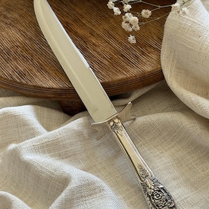 May include: A silver carving knife with an ornate handle. The blade is long and sharp, and the handle features a detailed floral design. The knife rests on a beige linen cloth.
