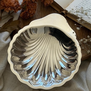 May include: A silver-colored, shell-shaped dish with a scalloped edge. The dish has a reflective surface and a detailed, ribbed design. The dish is likely made of metal and is suitable for serving or display.