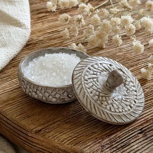 May include: A small, open ceramic bowl filled with coarse white salt, next to its lid. The lid and bowl have a textured, carved design. The items sit on a wooden surface, with dried white flowers in the background. The bowl is likely used for storing salt.