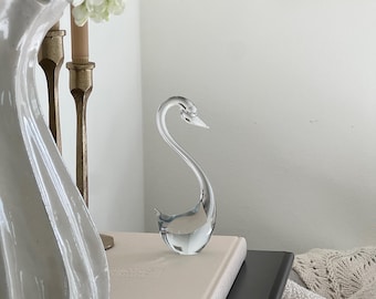 May include: A clear glass swan figurine with a curved neck and beak, resting on a light-colored book. The swan is positioned on a black surface, with a white vase and gold candle holders in the background.
