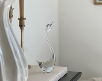 May include: A clear glass swan figurine with a curved neck and beak, resting on a white book. The swan is positioned on a black table, with a white vase and flowers in the background. The overall aesthetic is elegant and decorative.